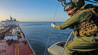 A still showing a military personnel on a helicopter above a large vessel. He is wearing green tactical gear and a helmet and gloves and is looking down at the boat. The water and sky are blue. 