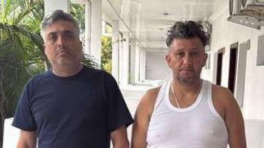 Carlos Alberto Rodelo, in blue T-shirt and shorts, stands next to Jorge Cubillos, in white vest and black trousers, with hands in pockets, on the balcony of their small hotel room in Kinshasa, with palm trees in the background.