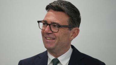 Andy Burnham looks off camera to the left. He is wearing a dark suit, white shirt and dark tie. 