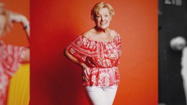 Old woman with short blonde hair wearing a red top and white trousers posing for a photoshoot 
