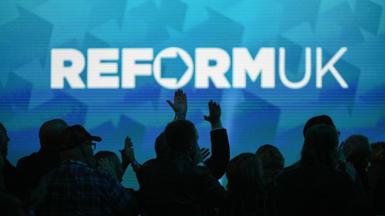 A Reform UK slogan projected on a wall with crowds pictured below in the dark.