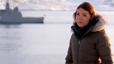 Katya Adler in a coat by the sea with a boat in the background.