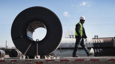 A man walking on a platform away from a huge role of steel that is taller than he is. He's wearing high-vis and a safety helmet.