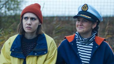 Two women sit next to each other outside. One is wearing a yellow rain mac with an orange beanie hat and the other is wearing a striped top with a blue jacket and sea captain's hat.