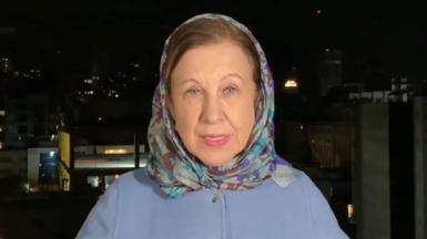 Lyse Doucet in a blue coat and headscarf, she is stood with a dark Tehran skyline behind her