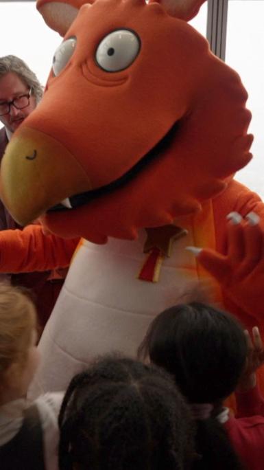 Person in orange dragon costume looks down at children in school uniform 