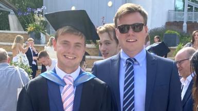 Jack and Ben O'Sullivan pictured at Jack's graduation. Jack is 23 and Ben is 27. They are white men with short, mousey brown hair. Jack is dressed in black graduation robes and a mortarboard, which he wears over a blue suit with pale blue shirt and pink tie. Ben also wears a blue suit, pale blue shirt and navy blue tie, with dark framed sunglasses. They're pictured outside on a sunny day around other graduates in the background.