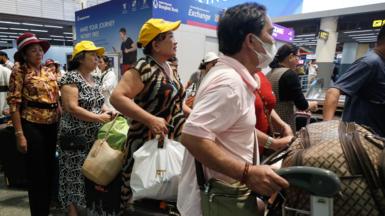Three women, two wearing yellow baseball caps, and a man holding a luggage trolley wait in Don Mueang International Airport in Bangkok, Thailand.
