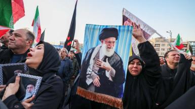 Protesters in Iran hold up flags and banners with Khamenei's picture on them. A woman in a black burqa hold her hand in the air and aother lady isclutching an image of Khamenei to her chest