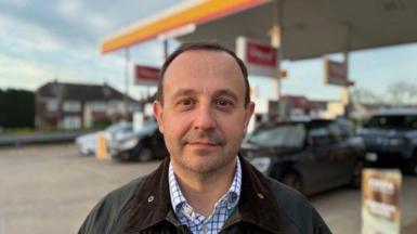Man with short hair in open necked shirt and Barbour style jacket standing on a petrol station forecourt