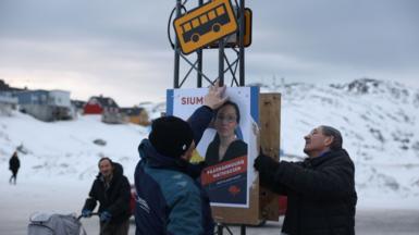 Two men work together to put up an election poster at a bus stop with snow on the ground around them