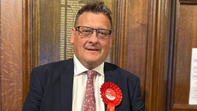 Karl Turner the succesful Labour candidate for Hull East pictured on election night in 2024 wearing a dark suit jacket, white shirt, and patterned pink tie, standing indoors against a wooden paneled wall. A large red rosette with the word ‘Labour’ and smaller text is pinned to the left side of the jacket. 