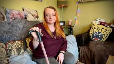 A woman in her 40s is sitting on a sofa in her living room holding a walking stick. She has long orange hair. Behind her is cushions and lighting.