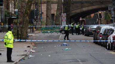 Police officers at the scene of an incident with a cordon in place in Friar Gate, Derby, on Sunday 29 March 2026. Debris remains on the road and cars are parked along the right.