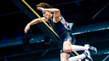 A man with medium length hair and wearing a blue vest and shorts is jumping over a horizontal yellow pole.