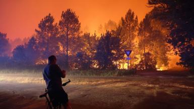 A person leans on a bike in front of a blaze that burns in a forested area