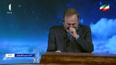Image shows a presenter on TV wearing a dark suit, with one hand to his face, eyes closed, and crying. The background behind him shows a blue sky with clouds