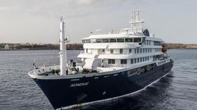 MV Hondius, a navy blue and white cruise ship, in the water. The sky is grey and the water is a dark blue, while in the distance you can see a strip of land. 