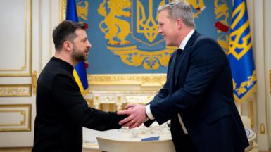 Volodymyr Zelensky, in black mockneck, puts hand forward, which Dan Driscoll, which grey hair and navy suit, grips in both hands. In the background are gold and blue insignia and flags