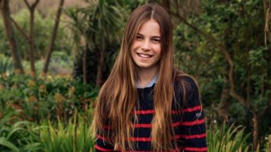 Photograph shows Charlotte wearing a black and red striped jumper and blue denim jeans, standing in a flowery field while smiling at the camera.