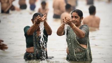 Two women wearing green sarees and gold bangles with their hair tied in a bun take a dip in the sacred waters of Sangam, the confluence of Ganges, Yamuna and mythical Saraswati rivers during the Maha Kumbh Mela festival in Prayagraj 