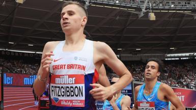 Robbie Fitzgibbon is running in a red, white and blue Great Britain vest with his name on the front, in front of a full crowd at an athletics stadium. He is in front over two other runners in blue vests