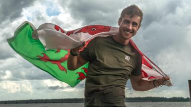 Ash Dykes is smiling at the camera after crossing the finish line from the Coppename river into the Atlantic. He is wearing an army green t-shirt and has a Welsh flag wrapped around his shoulders. The sea and Amazon rainforest are in the background and the sky is cloudy.