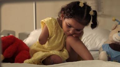A young girl wearing a yellow dress and with bunches in her hair rests her head on her knee while sitting on a bed surrounded by soft toys