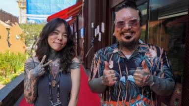 Kali (left) and Gil Taeho (right). Both have tattoos over the arms and are smiling at the camera
