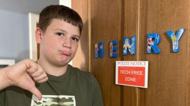 A teenage boy stands infront of door with the sign 'tech free zone' attached. He looks unhappy and is pointing downwards with his thumb.