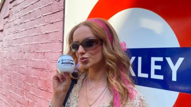 A young woman with long strawberry-blonde hair wears sunglasses and pouts at the camera, she is holding a white BuzzBallz container to her face. She is wearing sunglasses and a lace top with multiple necklaces on. Behind her is a London Transport Tube station sign and a pink brick wall.