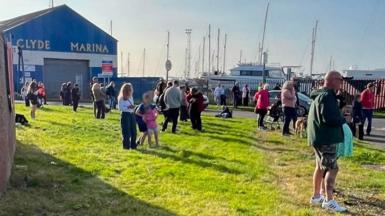 A crowd of people stands on a grassy area beside Clyde Marina, with yachts and sailboat masts visible behind a fence in the background. A large blue building marked “CLYDE MARINA” stands to the left, while people gather along the roadside and waterfront under bright sunshine. A speed limit sign and several parked vehicles are visible near the marina entrance.