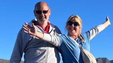 Andrew and Dawn Searle smile for the camera while standing outside - she is wearing a blue top with her arms stretched out wide, while he has a grey jacket/top on and is also smiling