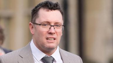 Image shows man in a light-brown suit with a silver tie and black-rimmed glasses. He has black hair and grey stuble.