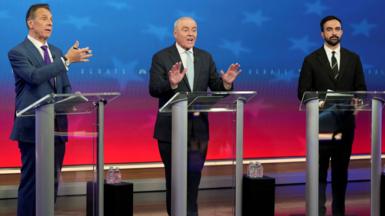 Andrew Cuomo, Curtis Sliwa and Zohran Mamdani stand at glass podiums on a stage with red and blue starred background and small stools with water next to them 
