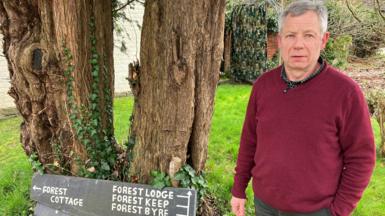 A man with short grey hair, wearing a burgundy jumper standing next to signage pointing to the holiday lets on his property