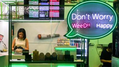 A brightly-lit neon shop window shows a woman in black gloves working surrounded by cannabis products