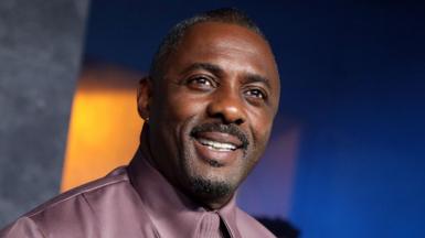 Idris Elba head and shoulders shot as he looks away from camera smiling, while wearing a purple shirt against a blue background, at the Luther premier in London in March 2023