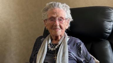 An elderly woman with short grey hair and bright eyes, smiles as she sits in a leather chair. She wears a navy shirt and grey scarf