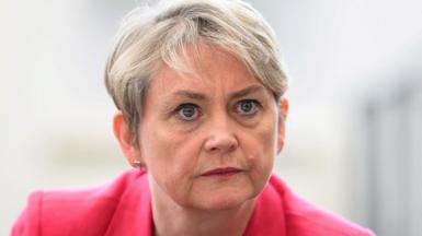 Home Secretary Yvette Cooper, a middle aged woman with short grey hair, stares intently at the camera. She is wearing a pink jacket