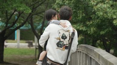 A man dressed up in a hoodie carries a baby in his arms while crossing a bridge in a park.