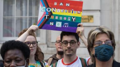A tightly cropped image of campaigners protesting for a ban on conversion practices. Two campaigners are in sharp focus, holding a sign featuring the progress pride flag and the words 'Ban Conversion Therapy Now' written in letters cut out from a newspaper or magazine. Both protestors are wearing glasses and have serious looks on their faces. Other protestors in the foreground are slightly out of focus.