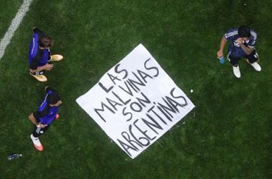 A banner saying "Las Malvinas Son Argentinas" is on the ground of a pitch with three players standing around it