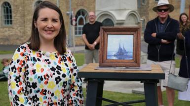 Theo Burrell wears a colourful top and stands next to a framed picture of a ship. She is standing on grass and people stand in the distance behind her 