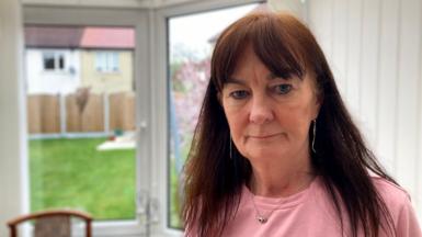 Chris Adamson, a woman with long brown hair, standing in a conservatory with a serious look on her face. She is wearing a pink top and necklace. A lawn, wooden fence and neighbouring houses can be seen through glass doors.