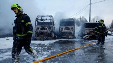 A firefighter walking in front of three burnt out semi-trucks with sleet and snow surrounding the vehicles