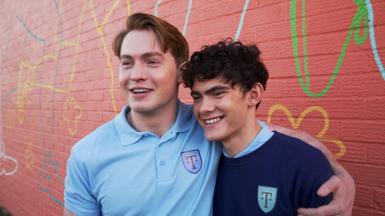 Actors Kit Connor and Joe Locke in their Truham High P.E kit from the TV show heartstoppers. Kit has his arm around Joe and they're both laughing at the camera.