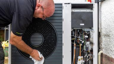 A male engineer wearing a black T-shirt wipes the outside of a black heat pump with a cloth. To the right of the picture the heat pump abuts the wall of a property with the pipes and electrical components visible