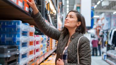 A woman with dark brown hair, wearing a green coat, shopping in a construction store. She is reaching up at a shelve which contains boxes.