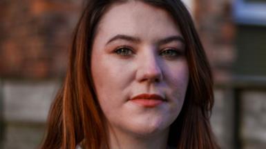 A still image of Jodie Morrow. She is staring at the camera, while she is stood against a wall outside. She has long, light coloured hair and is wearing a blue, knitted cardigan over a white t-shirt.